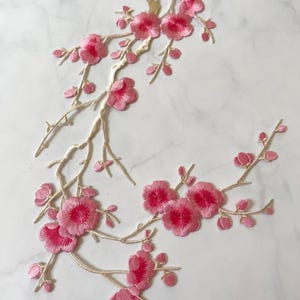 May include: Embroidered floral applique featuring pink cherry blossoms and buds on gold-colored branches. The blossoms have a darker pink center. The applique is likely for clothing or craft projects.