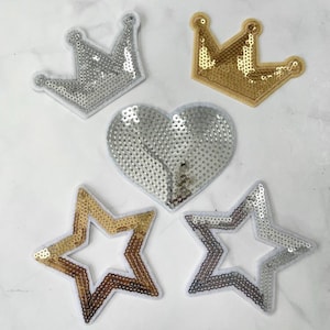 May include: Five iron-on patches with a sparkly, sequined finish. The patches include a gold crown, a silver crown, a silver heart, a gold star, and a silver star.