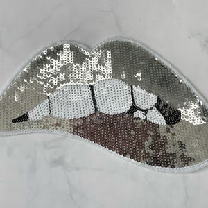 May include: A silver sequined lips patch with a white border. The lips feature black and white detailing, creating a glamorous design. This patch is suitable for clothing or accessories.