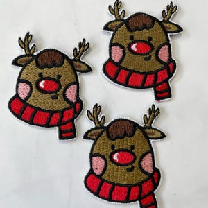May include: Three embroidered iron-on patches of a cartoon reindeer wearing a red and white striped scarf. The reindeer has brown fur, a red nose, and pink cheeks.