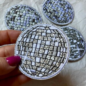 Op de afbeelding: Vier geborduurde patches in de vorm van discoballen. De patches hebben een mozaïek van zilveren en witte vierkanten, omlijnd met zilveren draad. Elke patch heeft een witte rand en een sterrenpatroon.