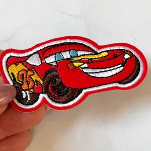 Può includere: Toppa ricamata raffigurante un'auto da corsa rossa dei cartoni animati con un volto sorridente, occhi blu e un design a fulmine giallo. La toppa ha un bordo bianco e pneumatici marroni. L'auto ha il numero 95 sul lato.