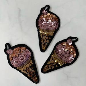 May include: Three pink and brown ice cream cone shaped patches with gold sequins. The patches are made of fabric and have a black outline.
