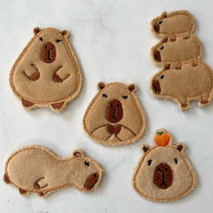 Może przedstawiać: Sześć brązowych filcowych naszywek w kształcie kapibary z haftowanymi detalami. Naszywki przedstawiają kapibary w różnych pozach, w tym jedną z małym pomarańczowym owocem na głowie i drugą z trzema kapibarami ułożonymi w stos.