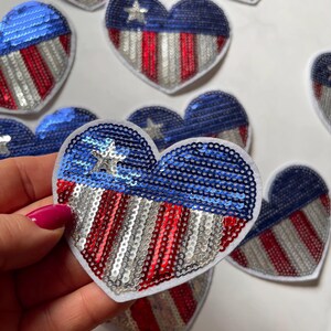 May include: Heart-shaped sequin patches in red, white, and blue, resembling the American flag. The patches feature a blue section with a white star and red and silver stripes. These decorative patches are ideal for patriotic crafts.