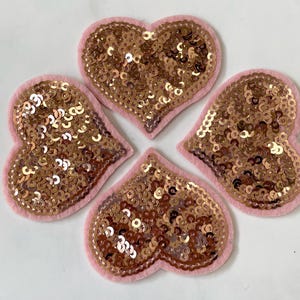 Peut inclure: Quatre patchs en forme de cœur en feutre rose avec des paillettes dorées. Les paillettes sont disposées de manière aléatoire.