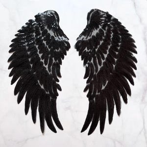 Op de afbeelding: Twee zwarte engelen vleugel applicaties met pailletten. Elke vleugel heeft een gedetailleerd veermotief met een doorschijnende, lichtgekleurde achtergrond. De pailletten vangen het licht op en creëren een glinsterend effect.