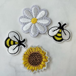 Puede incluir: Cuatro parches bordados para planchar que representan una margarita blanca, un girasol amarillo y dos abejas amarillas y negras.