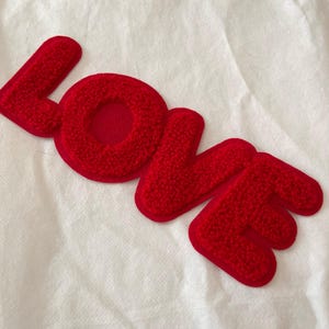 Puede incluir: Un parche rojo texturizado con la palabra "LOVE" en letras en relieve. Las letras están en una fuente audaz y juguetona, sobre un fondo blanco. El parche es probablemente para ropa o accesorios.