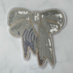 May include: A silver sequined bow with a white felt backing. The bow is a decorative embellishment for clothing or crafts.