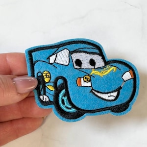 May include: A blue embroidered car patch with black outlines. The car has a smiling face, black eyes, and white teeth. The patch has a yellow lightning bolt design and the number 4 on the side.