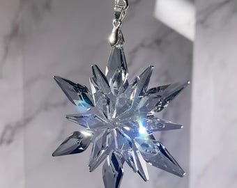 May include: A clear, faceted crystal snowflake pendant. The pendant is suspended from a silver-tone clasp and a strand of black beads. The snowflake design has sharp, pointed edges and reflects light, creating a sparkling effect.
