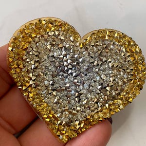 May include: A heart-shaped brooch covered in clear and gold rhinestones. The rhinestones are arranged in a gradient pattern, with the gold rhinestones on the outside and the clear rhinestones in the center.