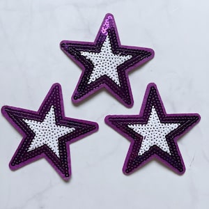 May include: Three purple star-shaped patches with white sequin centers. The stars have a layered design with a dark purple border and a lighter purple middle section. The sequins add a sparkly texture to the patches.