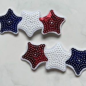 May include: Two sets of star-shaped patches. Each set has three stars in red, white, and blue sequins, with white felt backing. The stars are arranged in a row, evoking a patriotic theme.