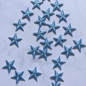 May include: A collection of light blue star-shaped embroidered patches. Each star has five points and is outlined with a darker blue thread. The patches are scattered on a white surface, suitable for crafting or embellishment.