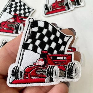 以下が含まれることがあります： 赤いレーシングカーとチェッカーフラッグが刺繍されたパッチ。車は赤で、黒い車輪と黒いドライバーが付いています。チェッカーフラッグは黒と白です。