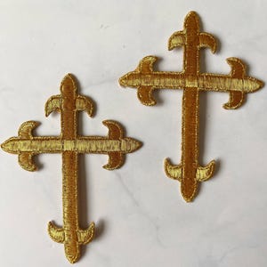 Puede incluir: Dos cruces doradas bordadas con un diseño de flor de lis. Las cruces están hechas de un hilo dorado brillante y texturizado y se muestran sobre una superficie blanca. Las cruces son idénticas en forma y tamaño.