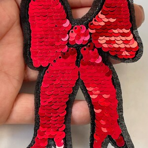 May include: A red sequined bow applique with a black fabric backing. The bow features a layered design with a central knot and two loops, all covered in shiny red sequins. This decorative embellishment is suitable for various craft projects.