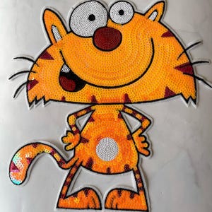 以下が含まれることがあります： オレンジと白のキラキラした猫のアップリケで、大きな笑顔と大きな目があります。猫の背中には白い斑点があり、ピンクとオレンジの虹色の尻尾があります。