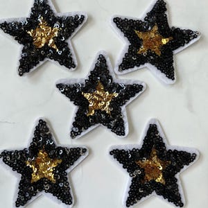 May include: Five black and gold star-shaped patches with sequins. The stars are on a white background.