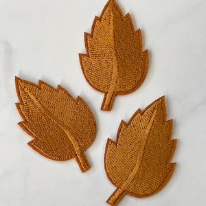 May include: Three embroidered fabric leaves in a warm orange color. The leaves are stitched with a detailed pattern and have a slightly raised texture.