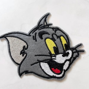 May include: A gray embroidered patch featuring the face of Tom from the Tom and Jerry cartoon. The patch has yellow eyes, a red mouth, and black whiskers and outlines. The patch is designed to be ironed on or sewn onto fabric.