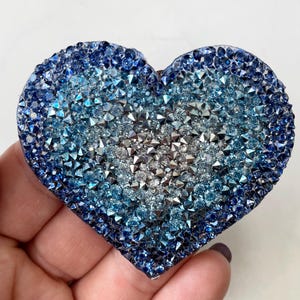 May include: A heart-shaped decorative object covered in blue and silver crystals. The crystals transition from dark blue at the edges to lighter blue and silver in the center. The object is held in a hand.