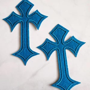 Może przedstawiać: Dwa niebieskie filcowe krzyże z dekoracyjnym wzorem. Każdy krzyż ma szczegółowy, wyszywany kontur w nieco ciemniejszym odcieniu niebieskiego. Krzyże umieszczone są na białej powierzchni.