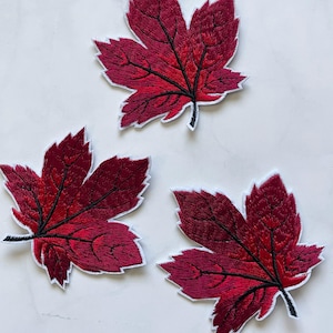 May include: Three red embroidered maple leaf patches. The leaves are a deep red color with black veins and a white outline.