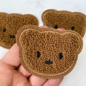 May include: Three brown fuzzy bear-shaped patches with embroidered black eyes and a mouth. The patches are sewn with a brown thread around the edges.