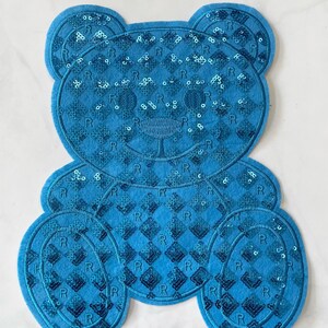 May include: A blue felt teddy bear applique with a checkered pattern and sequins. The bear has a simple face with a small smile.