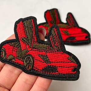 May include: Two red embroidered iron-on patches of a sports car with the doors open. The car is outlined in black and has green details.