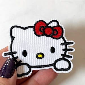 May include: A white embroidered Hello Kitty patch with a red bow and black outlines. The design features black eyes and whiskers. The patch is held by a hand with a purple nail. The patch is a popular character.