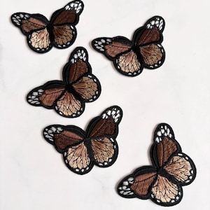 May include: Five embroidered butterfly patches. Each butterfly has brown and tan wings with black outlines and white accents. The patches are on a white surface.