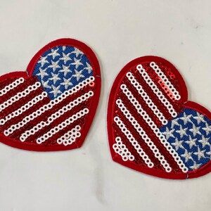 May include: Two red felt heart-shaped patches with white and blue sequins. The patches are decorated with the American flag design, one with red and white stripes and the other with blue stars.
