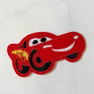 May include: A red embroidered patch featuring a cartoon car with black tires and a white smiling mouth. The car has black eyes and a yellow and orange flame design on the side. The patch is likely for clothing or accessories.