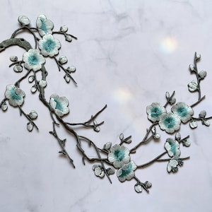 Könnte beinhalten: Gesticktes Blumenast-Design mit hellblauen und weißen Blüten mit türkisfarbenen Zentren. Die Äste sind dunkelgrau. Das Design befindet sich auf einem weißen Marmorhintergrund. Dieser dekorative Aufnäher ist ideal für Kleidung oder Bastelprojekte.
