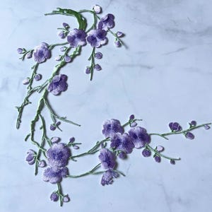 Peut inclure: Motif floral brodé avec des fleurs violettes et des tiges vertes. Les fleurs et les bourgeons délicats sont finement détaillés, créant un élément décoratif. Idéal pour embellir les vêtements, les accessoires ou les projets de décoration intérieure.