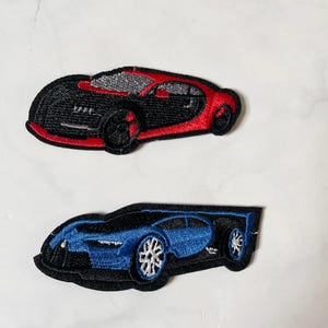 May include: Two embroidered car patches. One is black and red, the other is blue and black. The patches are in the shape of sports cars, with detailed stitching on the wheels and body. The patches are likely for clothing or accessories.