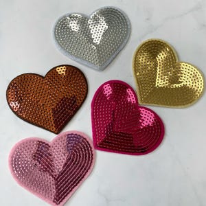 Puede incluir: Cinco parches en forma de corazón con adornos de lentejuelas en varios colores, incluyendo plata, oro, cobre, rosa y rojo.