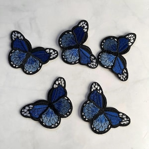 May include: Five embroidered butterfly patches. Each butterfly features a gradient of blue hues, from deep navy to lighter shades, with white accents on the wing edges. The patches have a black border, and are arranged on a white surface.