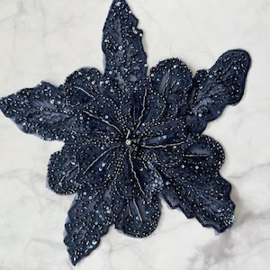 May include: A navy blue flower-shaped embellishment with sequins and beads. The embellishment is made of fabric and has a delicate, intricate design.