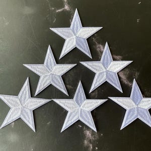 May include: Six white embroidered five-pointed stars with a light blue background. The stars are arranged in a triangular pattern.