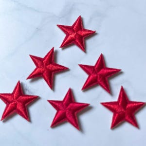Puede incluir: Seis parches de estrellas rojas bordadas sobre una superficie blanca. Cada estrella tiene cinco puntas y una apariencia texturizada, sugiriendo una construcción de tela. Ideales para manualidades o decoración.