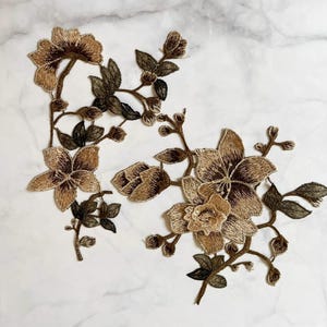 Puede incluir: Apliques florales bordados en tonos marrón y verde oliva. Los apliques presentan costuras detalladas, que representan flores, capullos y hojas. El diseño se encuentra sobre un fondo de mármol blanco, mostrando los intrincados detalles del bordado.