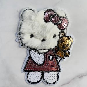 Könnte beinhalten: Ein weißer Plüsch-Patch mit einer Cartoon-Katze mit einer rosa Pailletten-Schleife und einem roten Pailletten-Kleid. Die Katze hält einen goldenen Pailletten-Teddybären. Der Patch ist mit schwarzen Perlen umrandet.