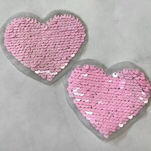Puede incluir: Dos parches en forma de corazón con lentejuelas rosas. Cada corazón está relleno de pequeñas lentejuelas rosas redondas y está colocado sobre un respaldo de malla gris. Los corazones son de color rosa claro y son ideales para adornar ropa o accesorios.