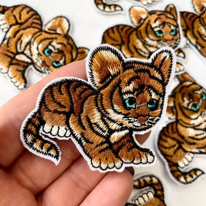 May include: A set of five embroidered iron-on patches featuring cute cartoon baby tigers with orange and black stripes. The patches are perfect for adding a touch of whimsy to clothing, bags, or other fabric items.