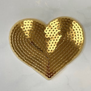 May include: A gold heart-shaped patch made of sequins. The sequins are arranged in a pattern that creates a swirling effect.